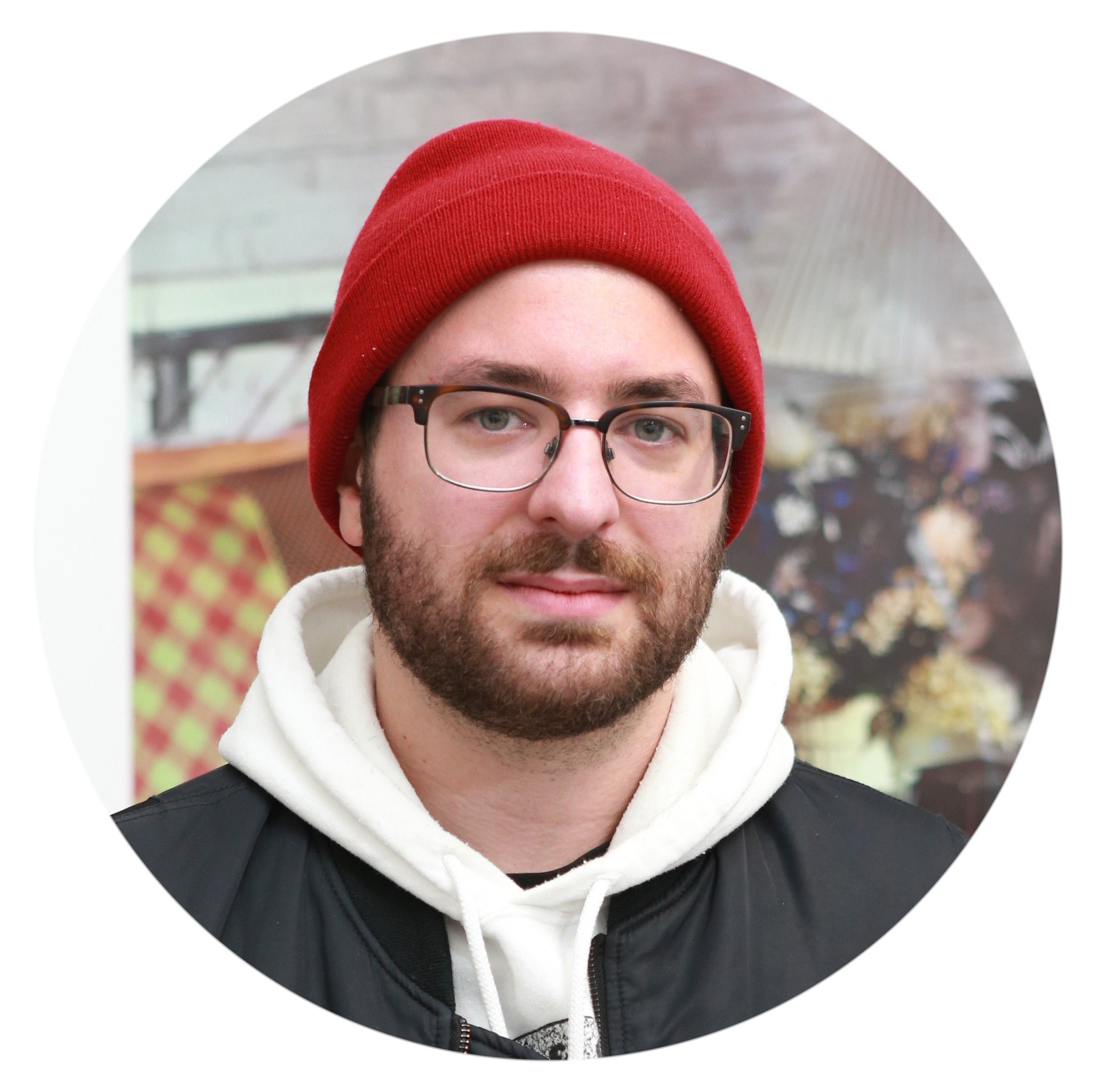 Headshot of a man with a brown beard wearing a red beanie and square glasses staring into the camera, wearing a black coat over a white hoodie.